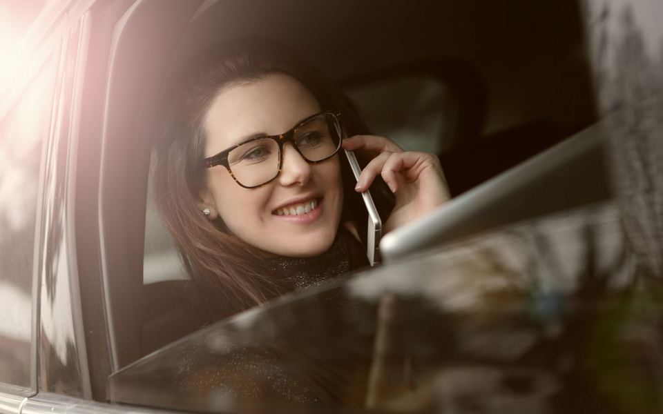 Smiling woman in glasses talking on her phone while sitting in a parked car